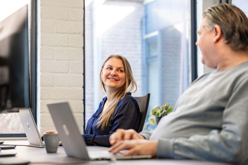 Twee mensen glimlachen zittend met laptops in kantoor.